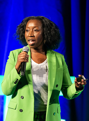 A black woman wearing lime green jacket is talking into a microphone. She is standing in front of a dark blue curtain